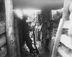 A member of the 1/7KR, stands in silhouette, while leaning on the wooden wall of a covered portion of a trench. Two additional soldiers stand in the background, in an uncovered part of the trench, almost washed out by the contrast.