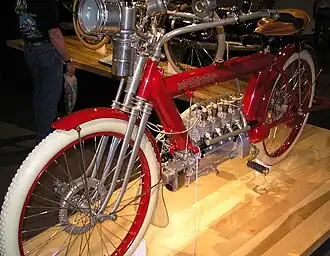 A bright red 1910 motorcycle with a flame headlamp, white rubber tires and a four-cylinder engine.