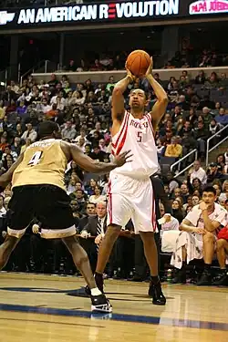 Howard is shooting a basketball over another Black athlete while an Asian athlete watches.