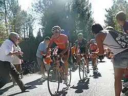 A line of three road racing cyclists, led by one in an orange jersey. Spectators line the road on either side, mere inches from the cyclists.
