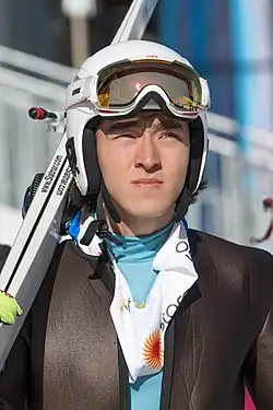 Colour photograph of Chingiz Rakparov aged 24, he is wearing full ski gear with his goggles on his helmet and skis over his right shoulder he is squinting looking towards the sun