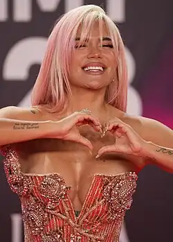 Karol G, wearing an ornate strapless dress, forms a heart with her two hands and smiles with letters behind her.