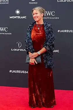 The image shows Kirsten Neuschäfer on the red carpet at the 25th Laureus World Sports Awards. The person is elegantly dressed in a floor-length red velvet dress and a dark blue patterned jacket, holding a grey clutch. She stands in front of a backdrop featuring the logos of Montblanc, IWC Schaffhausen, and the hashtag#LAUREUS24.The overall ambiance is one of luxury and celebration of sporting excellence.