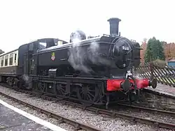 A black pannier tank locomotive is passing along a single track through woodland. The locomotive is pulling a train of a covered red wagon, three grey open wagons, and a red guard's van.