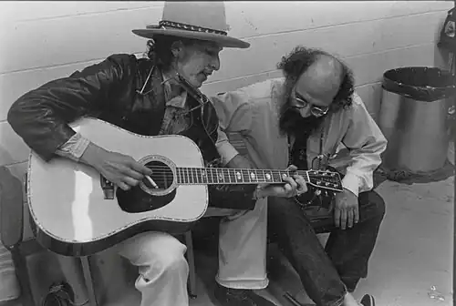 Dylan, wearing a hat and leather coat, plays guitar and sings, seated. Crouched next to him is a bearded man, listening to him with head bent.