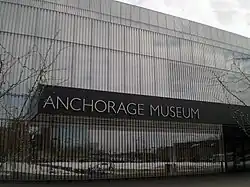 The mirrored facade of a building is seen with a stainless steel nameplate reading Anchorage Museum.