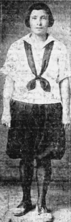 A young white woman with short dark hair, wearing a white middy blouse with short dark skirt or bloomers, and dark tights, with flat athletic shoes
