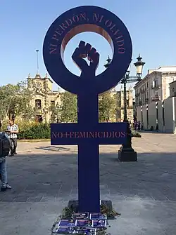 A symbol of Venus with a closed fist raised in the center. It has written the phrases "neither forgiving nor forgetting" and "no more femicides" in Spanish.