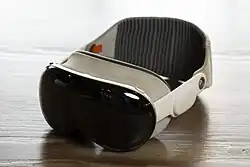 Photo of an Apple Vision Pro, on a stand, with a battery. A man stands in the background in a blue shirt.