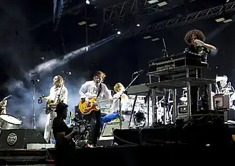 A side angle photo of a band playing in front of an audience under a soft white light. Several band members are in the photo. One is playing a guitar and holding a tambourine, one is playing a guitar, and one is playing an electric piano.