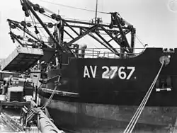 Black and white photo of a ship docked at a wharf. The ship's cranes are lifting a container above a truck.