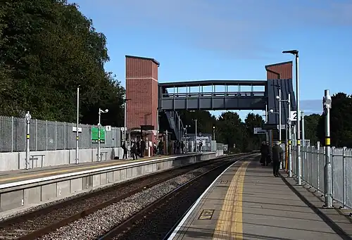 A railway station seen from the platform, with two tracks between the platforms and a footbridge over them with stairs and brick towers for lifts on each side.