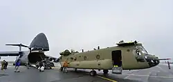 A green helicopter with its rotors removed in front of a large grey transport aircraft with its nose raised