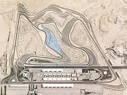 A tarmac motor circuit in a desert location, which is lined with concrete run-off areas, and features a large grandstand opposite the pit garages.
