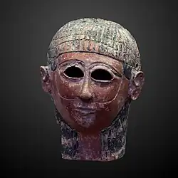 An ancient mask. The individual has brown skin, and black hair. There are holes at the eyes. Lines go down the face that are interpreted as a beard.