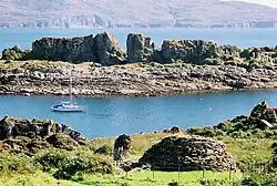 A rocky island with a small circular structure on it. In the middle distance there lies a yacht in blue water with another rocky isle beyond and a high shoreline in the distance.