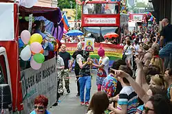 Three people in ski masks march in a LGBT Pride parade. One is holding a sign with a religious-style icon portrait of a member of the group.