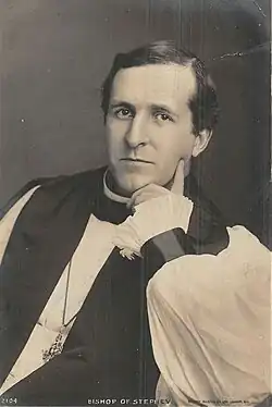 Black-and-white photo of a dark-haired white man, seated and resting his left hand around his chin, dressed in bishop's robes (rochet and chimere, pectoral cross and clerical shirt)