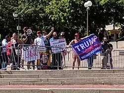 A group of people hold signs and flags, including a Trump 2020 flag, while standing against police barricades around Boston's Government Center.