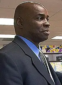 A man, wearing a black jacket with blue shirt, is standing in the middle of a classroom.