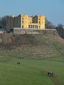 A yellow-painted house on a large stone terrace above a grass slope. It has two prominent octagonal towers at the corners with a single-storey arcade between them. Although most details are quite plain, overall it is an early example of the Jacobethan revival style.