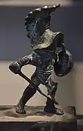 A statuette of a Hoplomachus (pl. hoplomachi) gladiator in ancient Rome, armed to resemble a Greek Hoplite (soldier with heavy armor and helmet, a round shield, a spear and a sword) wearing a bronze helmet, a manica on his right arm, loincloth (subligaculum).