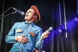 a man with a mustache, red beret and denim shirt sings into a microphone while holding a different microphone