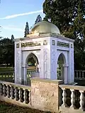 Roughly cubic white stone structure, topped by a gilded dome. Each of the four faces of the structure features a large pointed archway, and the two arches facing the camera also feature ornate Arabic calligraphy.