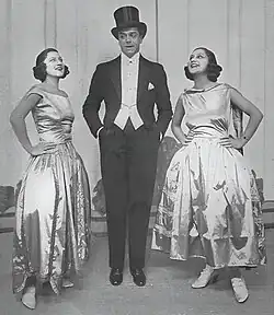 Tall, clean-shaven white man in top hat and tails with young white women in evening gowns on each side of him