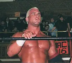 A white male standing in a wrestling ring with black ropes with his mouth open wearing wrestling gear in front of a crowd.