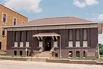 A midday film photo of the Carnegie Library Building in Carroll, Iowa. It is a brick building with Roman-style friezes under the windows depicting torches and scrolls. Major authors' names are carved in the upper part of the wall. The authors are Homer, Shakespeare, Dante, Milton, Longfellow, and Virgil. In the center above the door are the words "PUBLIC LIBRARY".