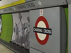 A platform wall: a panel contains the London Underground roundel with "CHARING CROSS" in the centre. A band contains the station name along the top of the wall. A decorative panel shows a photo-montage of Lord Nelson from the top of Nelson's column and the lions from around its base