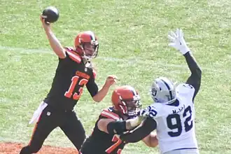 A photo of a quarterback in a brown #13 jersey throwing a pass while an opposing player attempts to block it
