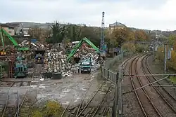 A scrapyard on the left with shunting engines and a lorry, with an active electrified railway line on the right