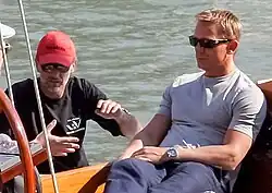 A man wearing a grey shirt, jeans and sunglasses sits on a boat at sea. Besides him, a man wearing a black shirt, red cap, and sunglasses talks to another, who is mostly off the picture.