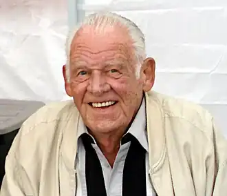 A man wearing a vanilla-colored jacket, with a white collared dress shirt and a black scarf around his neck