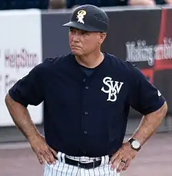 A man in a black baseball jersey with "SWB" written in white letters on the left breast pocket, a black coaches helmet with an "SWB" on the front, and white pants stands at third base lines.