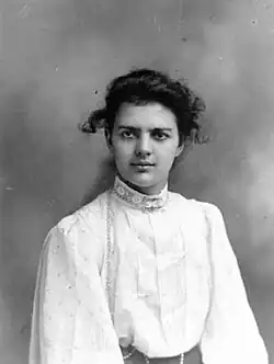 A black and white image of a woman facing forward looking just slightly to the left of the camera, she has dark hair, pulled back, but with a few curls of hair loose. She is wearing a high collared puffy white blouse.