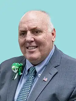Flynn is shown in a front-facing bust-length photograph. He is wearing a grey suit jacket, a blue button-up shirt, and a blue tie tightly-dotted with a pattern depicting green four-leaf clovers in recognition of St. Patrick's Day. He is wearing a green corsage on his right lapel (shown on the lefthand side of the photograph), also in recognition of St. Patrick's Day. On his left lapel (which is shown on the righthand side of the photograph), he is wearing pin depicting the national flag of the United States. Flynn is an older caucasian male with shortly-cut grey hair which has partially-receded above his forehead.
