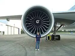 Aircraft engine, forward-facing view with a Boeing engineer in front to demonstrate the engine's size. The engine's large circular intake contains a central hub with a swirl mark, surrounded by multiple curved fan blades.
