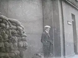 "Black and white photograph of O'Malley wearing a suit and tie and cap, with hands in pockets, looking partly away from the camera as he stands outside a tall building fortified by sandbags to the left"