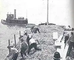 men shoveling shells into water from scow being towed by another boat