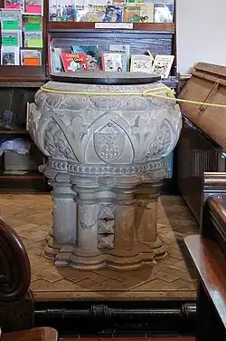 A wide, round-bodied font of pale stone with several rounded pillars forming its base. The stonework is elaborately carved. Around the top is a thin yellow rope, and a bookshelf stands in the background.