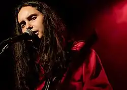 Francis of Delirium photographed from the shoulders up, singing into a microphone with her head tilted and her eyes mostly shut. She is wearing a red shirt in red lighting and holding an instrument that is difficult to identify. She has wavy dark brown hair that falls below her shoulders.