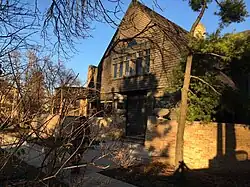 Exterior of the house as seen from Forest Avenue. There is a brick wall in front of the first story, where the entrance is located. The second story is within a triangular gable and has large windows.