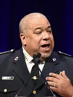 Harrison, in police uniform, speaking on a black-purple background