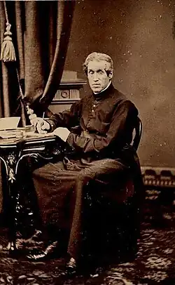 A man is pictured sitting down, facing the camera. His right hand is holding a pen to paper which sits on a table to the man's right, and his left hand rests on that table. The man is dressed in the clothes of a Christian religious leader.