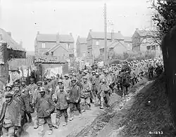 a large crowd of German soldiers walking along a small road under the supervision of soldiers wearing British equipment on horseback. Multiple houses can be seen in the background.