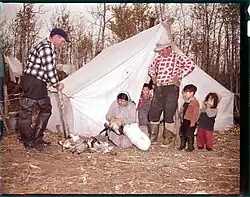 A man stands in the entrance of a tent with his hands on his hips, looking down smiling as a woman cleans some geese that have just been hunted. Children crowd around the man's legs, excited. Another man in waders leans on the pole of a tent nearby, also watching.