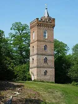 A colour photograph of a four-storey tower in red-brown brick with a crenelated parapet and a spire capping the staircase turret